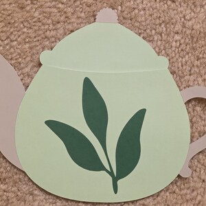 May include: A light green paper teapot cutout with a gray handle, spout, and lid. The teapot features a dark green leaf design on the front. The cutout is on a textured, tan surface.