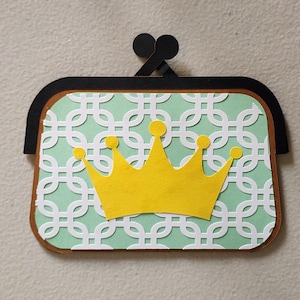 May include: A papercraft purse with a mint green background featuring a white geometric pattern. A yellow crown is centered on the purse. The purse has a brown border and a black clasp.