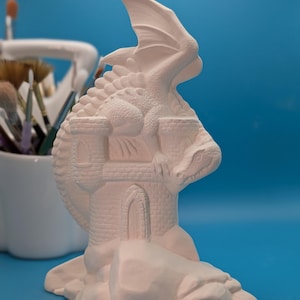 May include: A detailed, unpainted ceramic dragon figurine perched atop a castle tower. The dragon has textured scales, a large wing, and a long neck. The castle features a brick design and arched doorway. The figurine sits on a rocky base against a blue background.