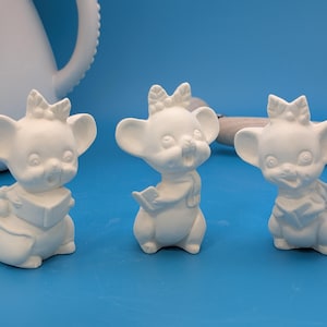 May include: Three unpainted ceramic mouse figurines with large ears, bows, and scarves. One mouse holds a book, another a book, and the third has a scarf. The figurines are on a blue surface.