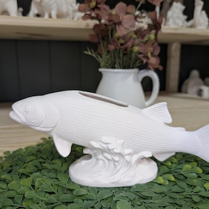 May include: A white ceramic fish figurine featuring a textured scale pattern on the body and a hollow opening on the top. The fish is mounted on a base sculpted to resemble splashing water, with detailed fins and a realistic head shape.