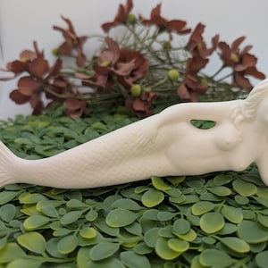 May include: A cream-colored mermaid figurine, reclining with one arm raised. The mermaid has a detailed tail and flowing hair. The figurine is resting on a bed of green leaves, with brown flowers in the background.