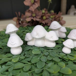 May include: A collection of white ceramic mushroom figurines with smiling faces. The mushrooms vary in size and are arranged on a bed of green leaves. The background includes a blurred arrangement of flowers.