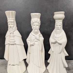 May include: Three white ceramic figurines of the Three Wise Men, each with detailed robes and headwear. The figures have long beards and are holding objects. The background is a solid dark gray.