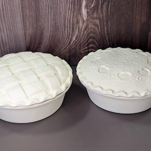 May include: Two white ceramic pie dishes. One has a woven lattice design on the top, and the other has heart and flower shapes. Both have a scalloped edge and sit on a gray surface.