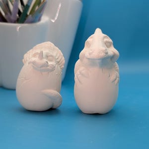 May include: Two unpainted ceramic dinosaur figurines. One is a dinosaur hatching from an egg, and the other is a dinosaur emerging from an egg. The figurines are white and sit on a blue surface. Paintbrushes are in a white container in the background.