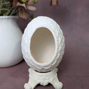 May include: A white, open-top egg-shaped decorative container with a detailed lace-like pattern. It sits on a matching white, ornate stand. The background includes a white vase with flowers.