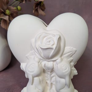 May include: A white ceramic heart-shaped vase with a rose and two bunny figures. The vase is unpainted and ready for customization. The design is suitable for home decor or as a gift.