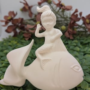 May include: A ceramic figurine of a mermaid riding a fish. The mermaid has her hair up and is waving. The fish has a large tail and fins. The figurine is off-white and sits on a bed of green leaves.