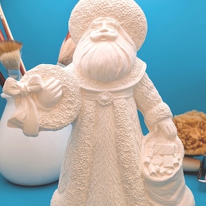 May include: A cream-colored Santa Claus figurine with a textured coat, hat, and beard. The figure holds a bag with a crest and a hat with a bow. Paintbrushes and a sponge are in the background.