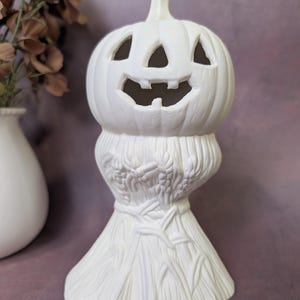 May include: A white ceramic pumpkin-shaped decoration with carved eyes, nose, and a smiling mouth. The pumpkin sits atop a detailed base resembling a bundle of wheat. Perfect for Halloween or fall decor.
