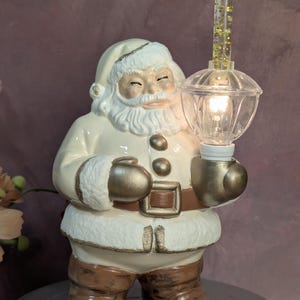 May include: A ceramic Santa Claus figurine in a cream-colored suit with gold accents, wearing brown boots and a belt. The figure holds a clear, illuminated lantern with a tall, glitter-filled top section, casting a warm light from its base.