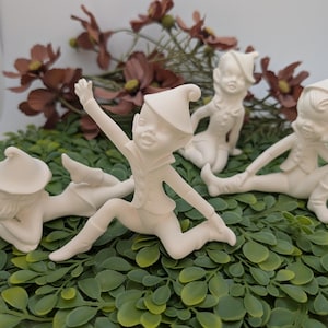 May include: Four off-white ceramic figurines of playful elves in various poses. One elf is leaping with an arm raised, while others are reclining or sitting. The figurines are set on a bed of green foliage, with brown flowers in the background.