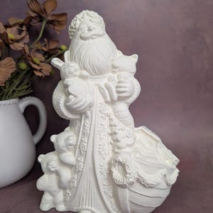 May include: A white ceramic Santa Claus figurine holding a rabbit and a cat, with three teddy bears at his feet. The figurine has a long white beard and is wearing a robe with floral detailing. A wreath is on the base.