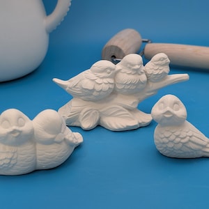 May include: A collection of unpainted ceramic bird figurines. The set includes a group of three birds perched on a branch, two birds nestled together, and a single bird. The birds are white and have detailed feather textures.