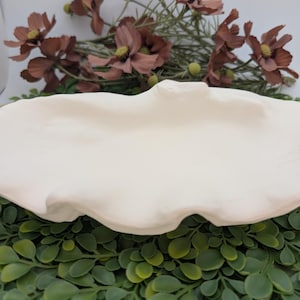 May include: A cream-colored, scalloped-edge ceramic serving dish. The dish is oval-shaped and sits on a bed of green foliage, with brown flowers in the background. The dish is likely used for serving food or as a decorative piece.
