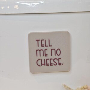 Könnte beinhalten: Ein weißer Behälter mit einem silbernen Griff und einem rechteckigen, hellfarbigen Aufkleber. Der Aufkleber trägt den Schriftzug "TELL ME NO CHEESE" in burgunderroten Buchstaben. Der Behälter ist teilweise sichtbar, mit einem Blick auf getrocknete Blumen unten.