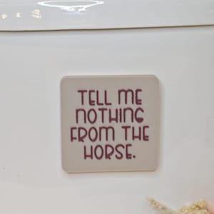 Könnte beinhalten: Ein quadratischer, hellbeige Magnet mit dem Schriftzug "TELL ME NOTHING FROM THE HORSE" in burgunderroter Schrift. Der Magnet ist auf einer weißen Oberfläche befestigt, mit einem Metallgriff oben.