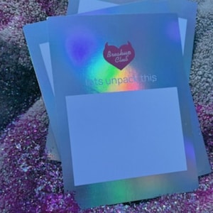 Könnte beinhalten: Ein Stapel schillernder Karten mit dem Text "Breakup Club" und "lets unpack this". Die Karten haben ein herzförmiges Logo und einen leeren weißen Bereich. Der Hintergrund ist eine glitzernde, mehrfarbige Oberfläche.
