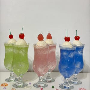 May include: Six colorful, layered drinks in clear, stemmed glasses. The drinks are green, pink, and blue, topped with whipped cream and a cherry or strawberry. Decorative candies are scattered around the base of the glasses.