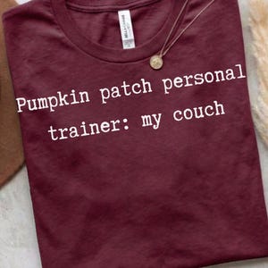 May include: A maroon short-sleeved t-shirt with a crew neckline and a white graphic print that reads: Pumpkin patch personal trainer: my couch. The shirt features a white brand label at the inner collar and a thin gold-toned necklace resting above.