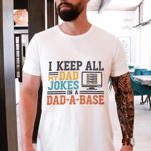 Könnte beinhalten: Ein weißes kurzärmeliges T-Shirt mit dem Text I KEEP ALL MY DAD JOKES IN A DAD-A-BASE in blauer, gelber und orangefarbener Schrift, begleitet von einer grafischen Darstellung eines Computermonitors mit einem Server-Rack-Symbol.