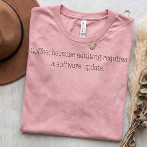 Könnte beinhalten: Ein altrosa T-Shirt mit kurzen Ärmeln und dem schwarzen Aufdruck: Coffee: because adulting requires a software update. Das Shirt hat einen Rundhalsausschnitt und ein weißes Markenetikett am Innenkragen, flach ausgebreitet dargestellt.