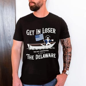 May include: A black short-sleeved t-shirt featuring a graphic of a figure in a boat holding a flag and a mug. The text reads: GET IN LOSER WE'RE CROSSING THE DELAWARE. The design includes a blue and white flag and a white boat on a black background.