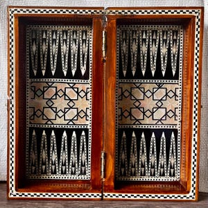 Könnte beinhalten: Ein Backgammon-Brett aus Holz mit Scharnierdesign. Das Brett hat einen dunkelbraunen Rahmen mit aufwendigen eingelegten Mustern in Weiß und Perlmutt. Die Spielfläche ist schwarz mit weißen und perlmuttfarbenen Details.