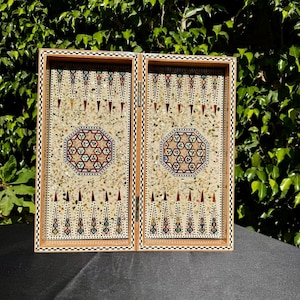 Op de afbeelding: Een houten backgammonbord met een decoratief ingelegd ontwerp. Het bord heeft een centraal achthoekig patroon en driehoekige punten in rood, groen en blauw. De lijst heeft een zwart-wit geblokte rand. Het bord is open en toont het speeloppervlak.