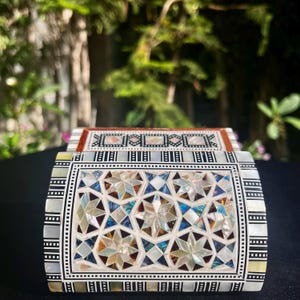 May include: A decorative box with intricate inlaid mother-of-pearl and geometric star patterns. The box features a rectangular shape with a hinged lid. The design incorporates a mix of white, iridescent, and brown tones, creating a visually appealing aesthetic.