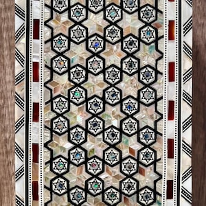 Pode incluir: Caixa decorativa retangular com um design de incrustação de madrepérola. A caixa apresenta um padrão geométrico de estrelas e hexágonos em preto e branco, com bordas vermelhas e pretas. O design interior tem detalhes iridescentes.