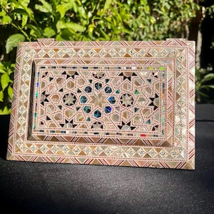 Puede incluir: Caja decorativa rectangular con incrustaciones de nácar. La caja presenta un patrón geométrico de estrellas y diamantes en colores iridiscentes, enmarcado por un borde con un diseño repetitivo. La caja está sobre una superficie oscura con follaje verde al fondo.