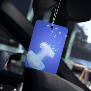 May include: A rectangular air freshener with a blue background and a white jellyfish illustration. The air freshener is hanging from a string, and the image is taken inside a car.