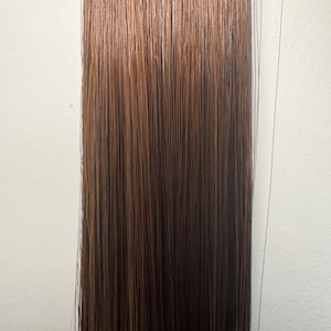May include: A bundle of hair extensions with a gradient effect. The top section is a light brown color, transitioning to a darker brown at the bottom. The hair strands are straight and sleek, with a natural shine.