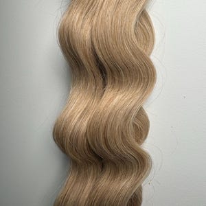 May include: Close-up of wavy, light brown hair extensions. The hair is a warm, golden blonde colour with subtle highlights. The strands are thick and appear to be of high quality, with a natural wave pattern. The hair is styled in a loose, flowing manner.