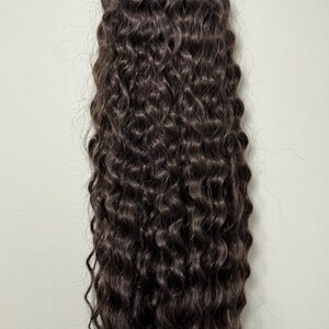 May include: A bundle of dark brown, tightly curled hair extensions. The hair has a deep, chocolate brown hue and a natural, wavy texture. The curls are uniform and tightly wound, giving a full and voluminous look. The hair is likely synthetic or human hair.