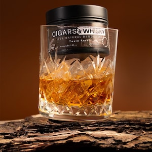 Op de afbeelding: Een zwarte cilindervormige houder met Cigars & Whisky 100% natuurlijke, gifvrije deodorant met een nettogewicht van 110 gram, geplaatst in een helder, gefacetteerd kristallen glas gevuld met amberkleurige vloeistof en ijsblokjes.