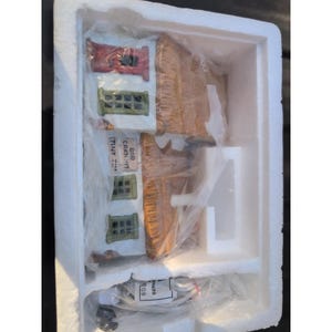 Puede incluir: Un juego de construcción de un pueblo en miniatura de cerámica, empaquetado en un contenedor de espuma de poliestireno. El edificio tiene un tejado rojo, ventanas verdes y el texto "TINY TIM" y "BOB CRATCHIT" en el lateral. El tejado es marrón y el edificio es blanco.