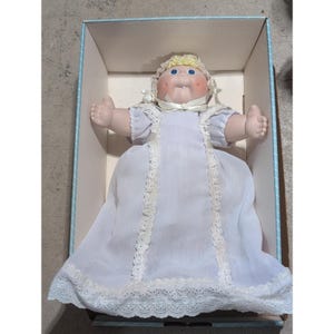 Op de afbeelding: Een pop met een zacht stoffen gezicht, blauwe ogen en blond wollen haar, gekleed in een witte muts en een lange witte jurk met kanten rand. De pop bevindt zich in een rechthoekige kartonnen doos met lichtblauw gestreepte randen.