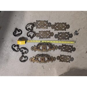 Puede incluir: Una colección de tiradores y manijas de cajones de latón antiguo en varios diseños ornamentados. Incluye varios tiradores ovalados, manijas decorativas y algunos tiradores curvos más pequeños. Una cinta métrica amarilla muestra las medidas en cm.