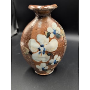 Puede incluir: Un jarrón de cerámica marrón con cuello estrecho y cuerpo redondeado. El jarrón está decorado con diseños florales blancos y azules. El jarrón tiene una superficie texturizada y un borde marrón oscuro.