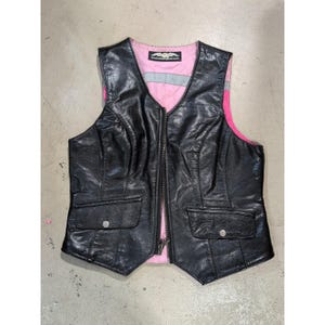 Puede incluir: Chaleco de cuero negro con forro rosa y detalles plateados. El chaleco tiene cuello en V, cierre de cremallera y dos bolsillos delanteros con cierres de botón a presión. Estilo motero clásico.