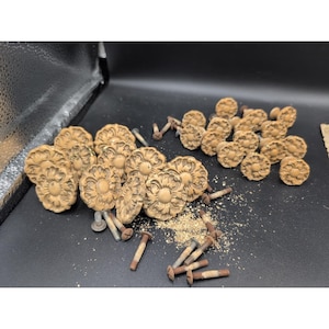 May include: A collection of vintage, light brown, floral-shaped cabinet knobs with matching screws. The knobs are arranged on a dark surface, with some screws and debris scattered around. The knobs have a detailed, carved design.