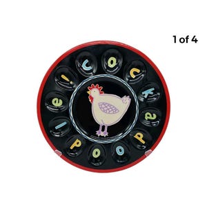 May include: A round, black ceramic egg holder with a red rim. The center features a cream-colored chicken illustration. The words "cock a doodle poo" are spelled out around the edge in colorful letters. The holder has 12 egg-shaped indentations.
