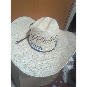 Op de afbeelding: Een lichtbeige strooien cowboyhoed met een brede rand en een geventileerde kroon. Een donkerbruine hoedband omcirkelt de basis, voorzien van een rechthoekige patch met de tekst 'The Cow Lot' en een kleinere ronde patch aan de zijkant. Een dun bruin koord is bevestigd.