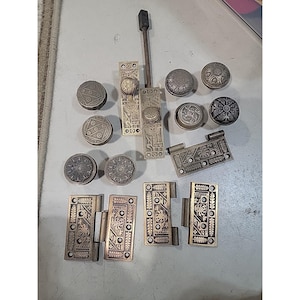 May include: An assortment of antique metal hardware, including door knobs, hinges, and a door latch. The hardware features intricate embossed designs and a tarnished finish, suggesting age and use. The pieces are arranged on a light gray surface.