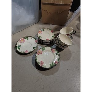 Op de afbeelding: Een set vintage kopjes en schotels. De witte keramische schotels en kopjes hebben een bloemenpatroon met roze bloemen en groene bladeren. De randen van de schotels en kopjes hebben een donkere rand. De set bevat meerdere schotels en kopjes.