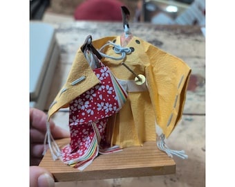 May include: A small, handcrafted Japanese doll, likely a geisha, made of paper and fabric. The doll features a yellow kimono with a red floral patterned under-kimono. It is mounted on a small wooden base and has black hair and a decorative headpiece.