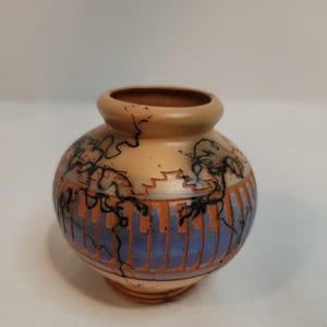 Könnte beinhalten: Kleine, runde Keramikvase mit hellbrauner Außenseite und brauner Innenseite. Die Vase hat ein blau-oranges Streifenmuster am Boden und schwarze Linienkunst. Der Rand ist dunkler braun.